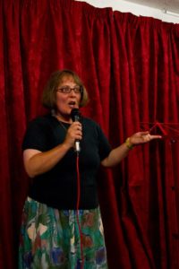 Susan performs her poetry on stage in front of a red curtain. She holds a microphone in her right hand and looks very animated.