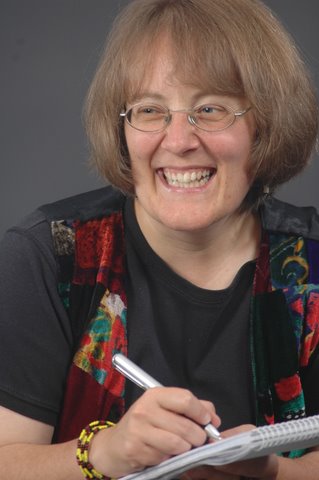 Close-up image of Susan smiling broadly as she writes in a notebook. She wears a short-sleeved black top and a colourful, patterned waistcoat.