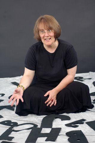 Susan sits cross-legged on the floor on a white sheet surrounded by big black cut-out letters. 