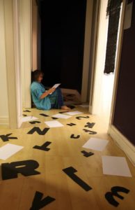 A distant view of Susan sitting on a shiny wooden floor surrounded by pieces of paper and big black cut-out letters.