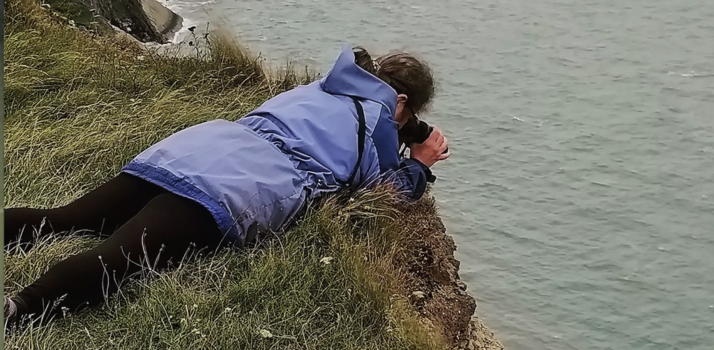 Susan, wearing a blue waterproof coat and black leggings, lies on her belly on the precipitous edge of a grass-topped cliff. She's using binoculars to look down at the sea several hundred feet below.